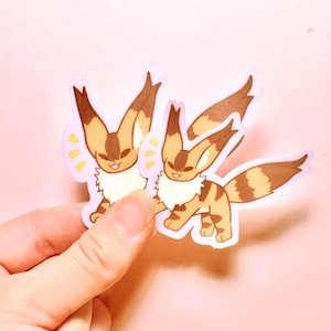 May include: Two die-cut stickers featuring a cartoon creature with large pointed ears, a fluffy white chest, and tan fur with brown stripes. The creature has a smiling expression and yellow motion lines near its head, rendered in a flat art style.