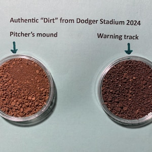 May include: Two small glass jars filled with dirt from Dodger Stadium. The jar on the left is labeled "Pitcher's mound" and the jar on the right is labeled "Warning track".