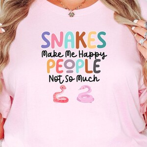 May include: Pink t-shirt with the text "Snakes Make Me Happy People Not So Much" in colorful letters. Two cartoon snake illustrations are below the text. The word "Pink" is in a pink rectangle in the bottom right corner.