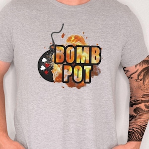 May include: Athletic Heather t-shirt with a graphic that says "BOMB POT" in orange and yellow. The design includes a bomb with a black casing and a red fuse, with a fiery explosion in the background. The shirt is a light gray color.