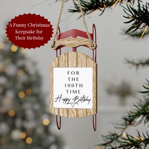 May include: A decorative sled ornament featuring a red metal frame and a light wood-grain base. A white rectangular plaque is centered on the wood, displaying the black text: FOR THE 100TH TIME Happy Birthday DAD with a small red heart.