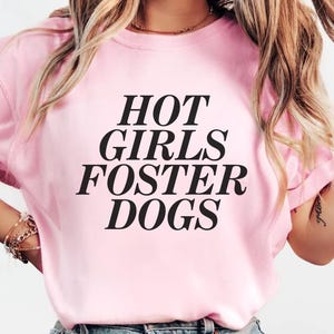 Könnte beinhalten: Hellrosa T-Shirt mit dem schwarzen Text "HOT GIRLS FOSTER DOGS". Das Oberteil hat kurze Ärmel und wird mit einer blauen Jeans getragen. Das Wort "Blossom" befindet sich oben links.