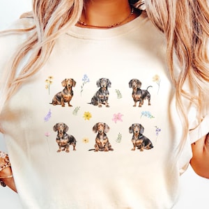 May include: Ivory-colored sweatshirt featuring a watercolor design of several dachshund dogs and colorful flowers. The dogs are depicted in various poses. The word "Ivory" is printed in the upper left corner.