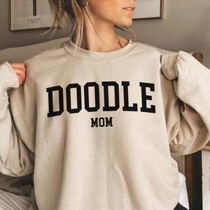 Może przedstawiać: Beżowa bluza z nadrukiem "DOODLE MOM" w kolorze czarnym.