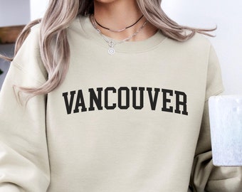 May include: A beige sweatshirt with the word "VANCOUVER" printed in black on the front. The sweatshirt is being worn by a person holding a white mug.