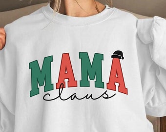 Op de afbeelding: Wit sweatshirt met de tekst "MAMA claus" in groene, rode en zwarte letters. Het woord "MAMA" staat in grote, vetgedrukte letters met een kleine kerstmanmuts boven de "A".