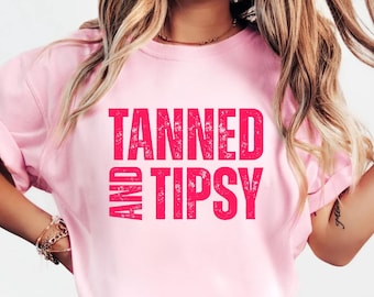 May include: A light pink t-shirt with the words "TANNED AND TIPSY" printed in bright pink. The shirt has short sleeves and a crew neck. The model is wearing blue jeans.