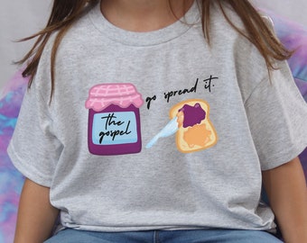 May include: A child wearing a grey t-shirt with a graphic of a jar of jam labeled "The Gospel" and a slice of bread with jam on it. The text "go spread it." is written above the graphic. The child is sitting on a colorful beanbag chair.