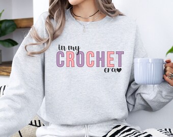 Puede incluir: Una sudadera gris con el texto "in my crochet era" en letras rosa y morado con un pequeño corazón negro.