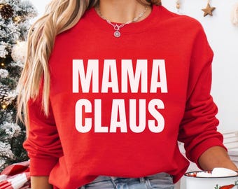 Op de afbeelding: Een rode trui met de witte tekst "MAMA CLAUS". De persoon houdt een witte mok vast met een illustratie van de kerstman. De achtergrond bevat een kerstboom en andere feestelijke decoraties.