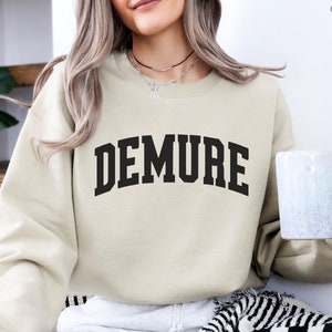 以下が含まれることがあります： 前面に黒い文字で「DEMURE」とプリントされたベージュのトレーナー。トレーナーは白いマグカップを持った人が着ています。