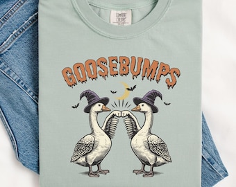 May include: A light blue t-shirt with a graphic of two geese wearing witch hats and holding up their fists. The text "GOOSEBUMPS" is above the geese in orange and black.