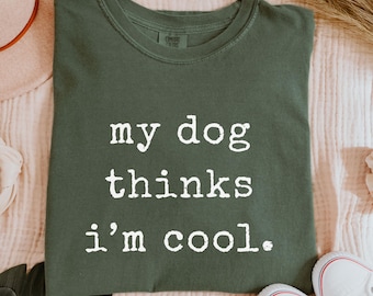 May include: A moss green t-shirt with the text "my dog thinks I'm cool" in white. The shirt is displayed on a pink surface with a hat, shoes, and other accessories.