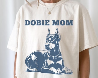 Può includere: T-shirt color avorio con la scritta blu "DOBIE MOM". Un'illustrazione blu di un cane Dobermann Pinscher è sotto il testo. Il cane è sdraiato, con un collare.