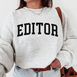 Pode incluir: Uma camisa de moletom cinza com a palavra "EDITOR" impressa em letras pretas.