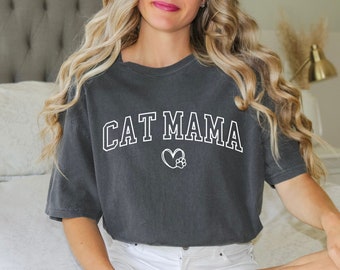 Könnte beinhalten: Eine Frau trägt ein dunkelgraues T-Shirt mit dem Text "CAT MAMA" in weißen Buchstaben und einem weißen Herz mit einem Pfotenabdruck darin. Das Shirt ist locker geschnitten und hat kurze Ärmel.