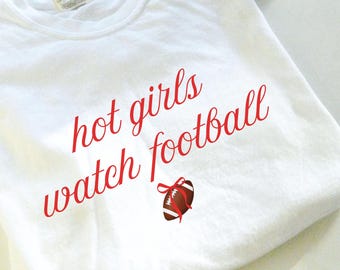 Puede incluir: Una camiseta blanca con el texto rojo "hot girls watch football" y un gráfico de fútbol americano. La camiseta está hecha de un material suave, ideal para uso casual.