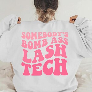 Peut inclure: Sweat-shirt blanc avec un texte rose qui dit "Somebody's Bomb Ass Lash Tech" dans une police rétro.