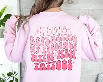 May include: Pink sweatshirt with the text "I love replacing my feelings with new tattoos" in a retro font.