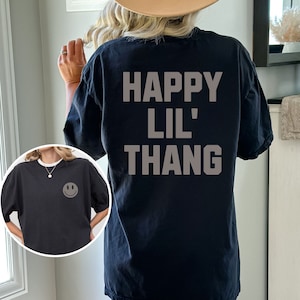 May include: Black t-shirt with the text "HAPPY LIL' THANG" in gray block letters on the back. A smaller image shows the front of the shirt with a smiley face graphic. The shirt is a casual, oversized style.