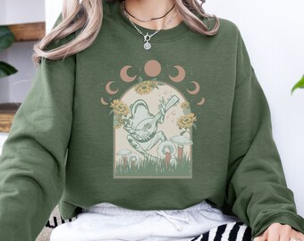 May include: A green sweatshirt with a graphic of a frog playing a guitar in a moonlit forest. The frog is surrounded by sunflowers and mushrooms. The text "Moonlit Melodies" is printed on the sweatshirt.
