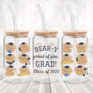 May include: Three clear glass tumblers with bamboo lids and glass straws. Each tumbler features a graduation-themed design with cartoon bears wearing graduation caps. The text "BEAR-Y proud of you, GRAD! Class of 2025" is printed on one tumbler.