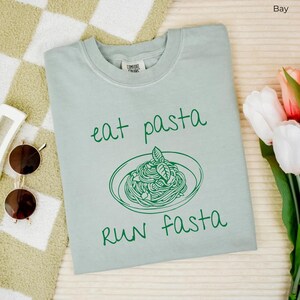Puede incluir: Una camiseta azul claro con un gr&aacute;fico verde de un plato de pasta y el texto "eat pasta run fasta".