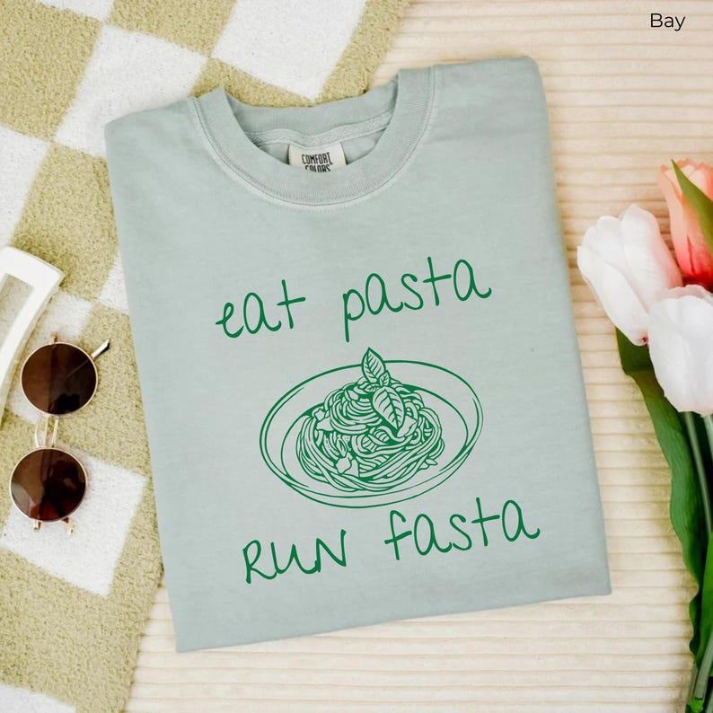Puede incluir: Una camiseta azul claro con un gr&aacute;fico verde de un plato de pasta y el texto "eat pasta run fasta".