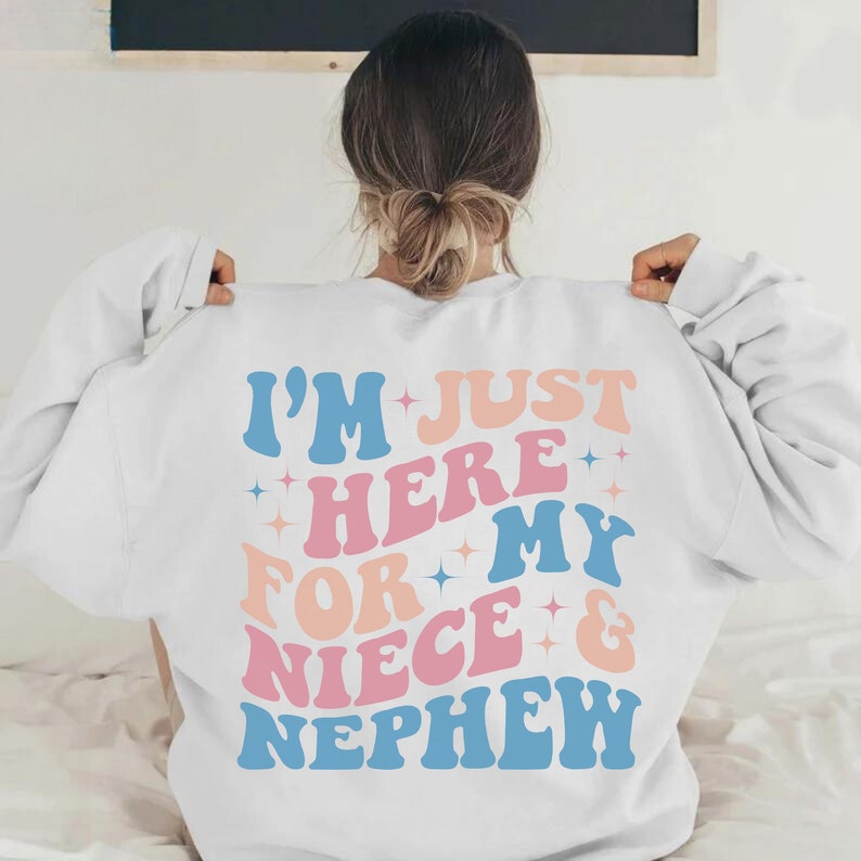Pode incluir: Camisola branca com o texto "I'M JUST HERE FOR MY NIECE & NEPHEW" em letras azuis, rosa e p&ecirc;ssego. O design inclui detalhes de estrelas. Uma camisola de estilo casual e confort&aacute;vel.