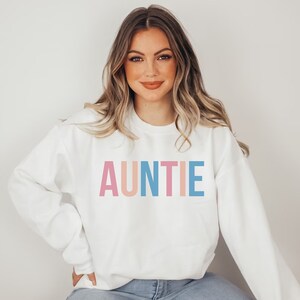 Pode incluir: Camisola branca com a palavra "AUNTIE" em letras de bloco rosa pastel, p&ecirc;ssego e azul. A camisola &eacute; de gola redonda e tem mangas compridas. A pessoa est&aacute; a usar cal&ccedil;as de ganga azuis.