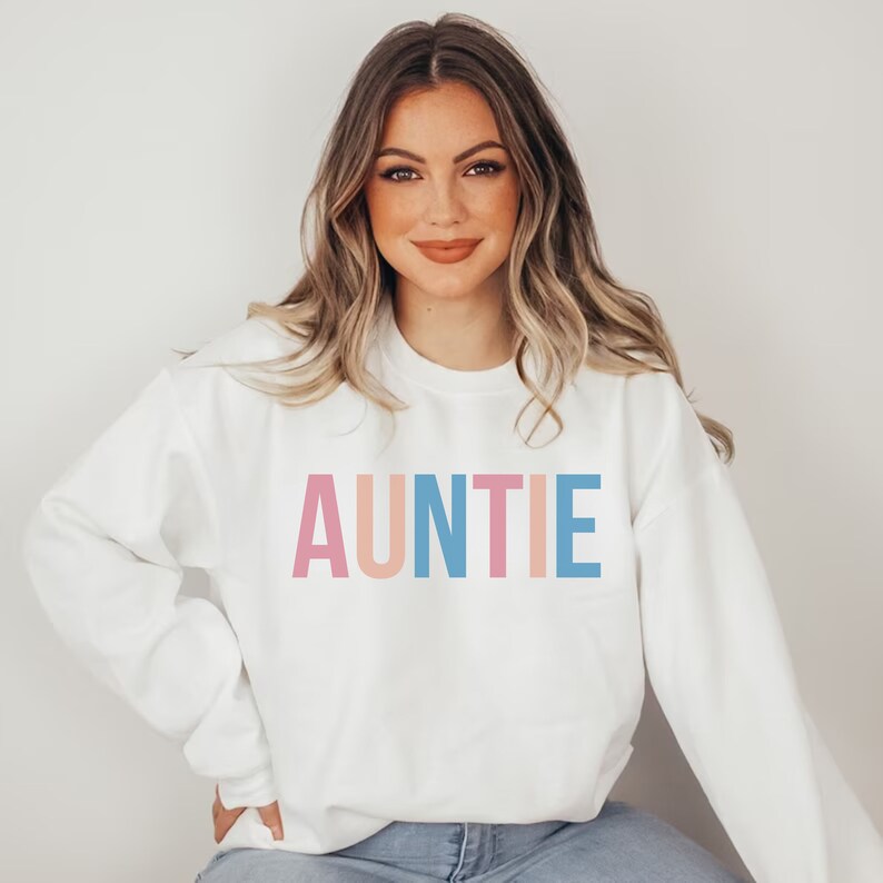 Pode incluir: Camisola branca com a palavra "AUNTIE" em letras de bloco rosa pastel, p&ecirc;ssego e azul. A camisola &eacute; de gola redonda e tem mangas compridas. A pessoa est&aacute; a usar cal&ccedil;as de ganga azuis.