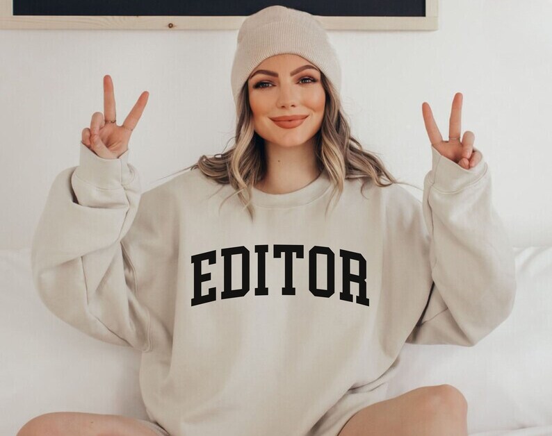 Pode incluir: Uma camisola bege com a palavra "EDITOR" impressa a preto na frente. A camisola tem gola redonda e mangas compridas. A modelo est&aacute; a usar um gorro branco.