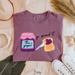 May include: Purple t-shirt with a graphic of a jar of jam labeled "The Gospel" and a slice of bread with jam on it. The text "go spread it." is above the bread.