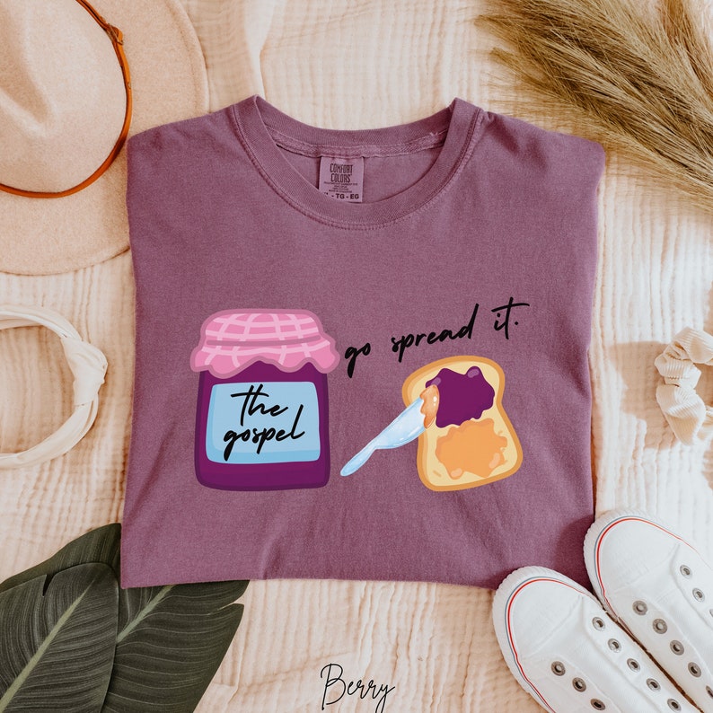 May include: Purple t-shirt with a graphic of a jar of jam labeled "The Gospel" and a slice of bread with jam on it. The text "go spread it." is above the bread.