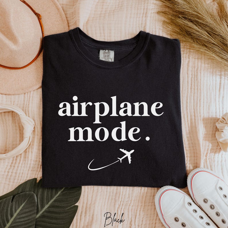 Op de afbeelding: Zwart T-shirt met witte tekst die "airplane mode" zegt. Naast de tekst staat een wit vliegtuigpictogram.