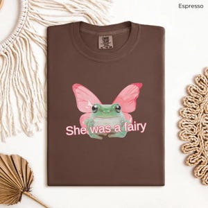Op de afbeelding: Een bruin T-shirt met een groene kikker die roze vlinder vleugels draagt. De tekst "She was a fairy" is in roze onder de kikker gedrukt.