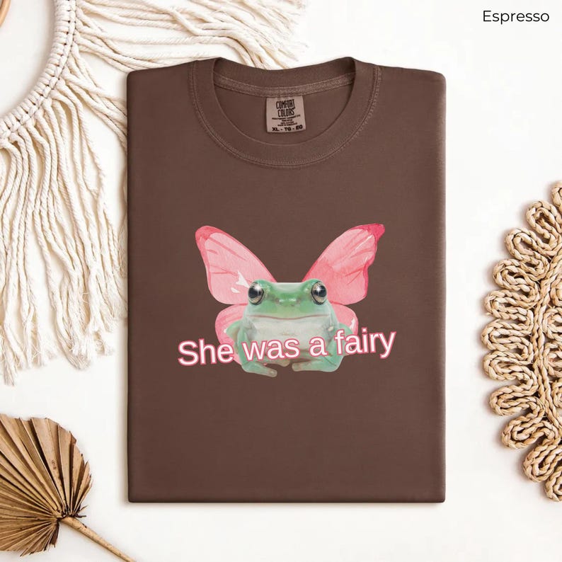 Op de afbeelding: Een bruin T-shirt met een groene kikker die roze vlinder vleugels draagt. De tekst "She was a fairy" is in roze onder de kikker gedrukt.
