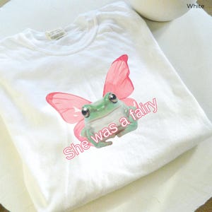 Op de afbeelding: Wit T-shirt met een groene kikker met roze vlinder vleugels. De tekst "She was a fairy" is op het T-shirt gedrukt.