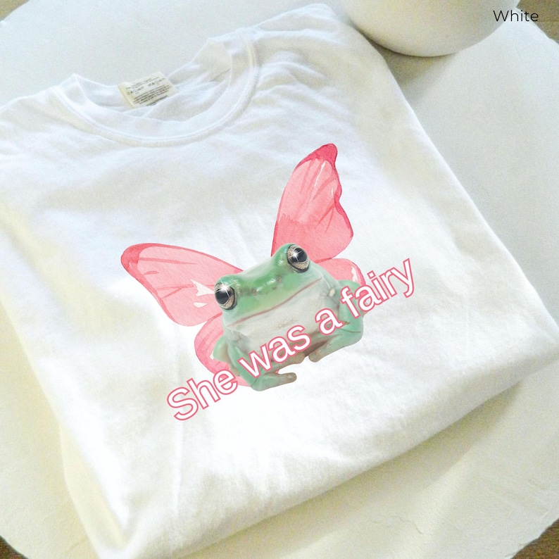 Op de afbeelding: Wit T-shirt met een groene kikker met roze vlinder vleugels. De tekst "She was a fairy" is op het T-shirt gedrukt.