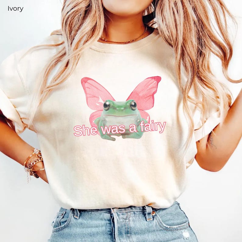 Op de afbeelding: Een wit T-shirt met een groene kikker die roze vlinder vleugels draagt. De tekst "She was a fairy" is in roze onder de kikker gedrukt.