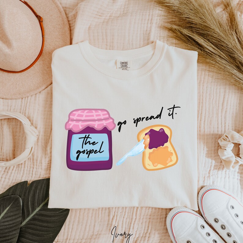May include: White t-shirt with a purple jar of jam that says "the gospel" and a slice of toast with purple jam on it. The text says "go spread it."