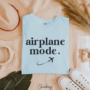Op de afbeelding: Een lichtblauw t-shirt met de tekst "airplane mode." in zwarte letters. Een klein vliegtuigpictogram vliegt boven de tekst.
