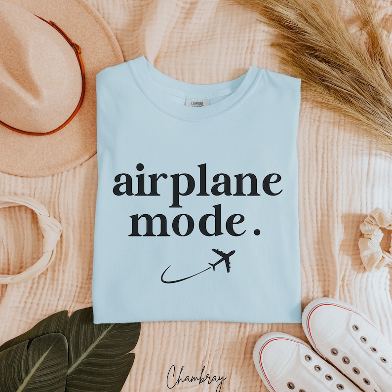 Op de afbeelding: Een lichtblauw t-shirt met de tekst "airplane mode." in zwarte letters. Een klein vliegtuigpictogram vliegt boven de tekst.
