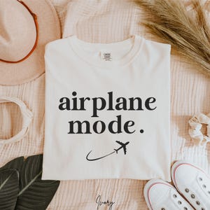 Op de afbeelding: Wit T-shirt met de tekst "airplane mode." in zwarte letters en een kleine vliegtuigafbeelding.
