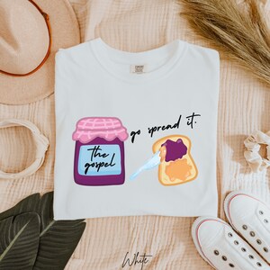 May include: White t-shirt with a graphic of a jar of jam labeled "the gospel" and a slice of toast with jam spread on it. The text "go spread it." is written above the toast.