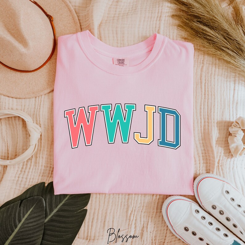Op de afbeelding: Roze T-shirt met de tekst "WWJD" in kleurrijke blokletters. De letters zijn omrand met zwart.