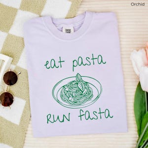 Puede incluir: Una camiseta de color morado claro con el texto "eat pasta run fasta" en verde. Un dibujo de un plato de pasta con albahaca est&aacute; en el centro de la camiseta.