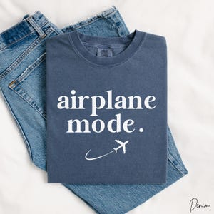 Op de afbeelding: Een donkerblauw t-shirt met de tekst "airplane mode." in witte letters. Een wit vliegtuigpictogram vliegt boven een witte gebogen lijn.