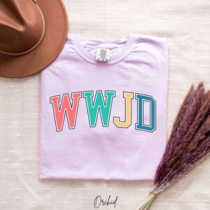 Op de afbeelding: Een lichtpaarse t-shirt met de tekst "WWJD" in kleurrijke blokletters. De letters zijn zwart omrand en elke letter is een andere kleur: roze, turquoise, geel en blauw.
