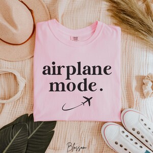 Op de afbeelding: Roze T-shirt met zwarte tekst die "vliegtuigmodus" zegt. Naast de tekst staat een klein vliegtuigpictogram.